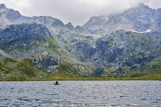View of a person rowing a small boat on the calm waters of the Suretta Seen near Splugen, surrounded by the rugged, rocky peaks of the Alps in Rheinwald, Grisons, Switzerland.