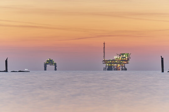 View of offshore oil and gas platforms illuminated at dusk on the calm sea with birds perched on wooden posts in the foreground in Lido di Dante, Emilia-Romagna, Italy.