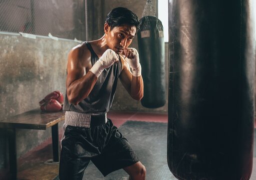 Focused young Asian muscular male boxer training with a heavy punching bag in a dark gym setting or fitness center, showing intense determination and sweat during a tough fighting workout session.