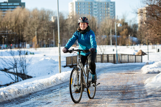 A happy mid-adult woman rides a bicycle on an icy path through a snowy winter park in Helsinki, Finland, enjoying active outdoor commuting.