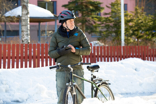 A senior man checks his smartphone near his bicycle on a snowy park road in Helsinki, Finland, navigating his winter commute