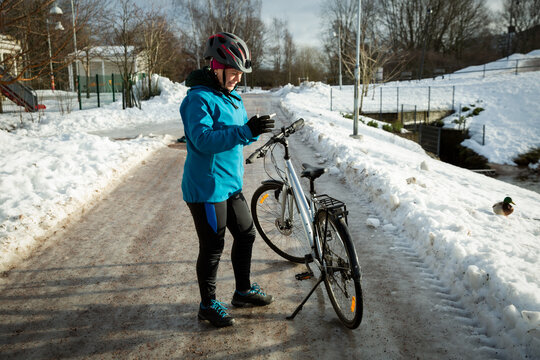 An active mid-adult woman checks her smartphone beside a bicycle on a snowy winter park road, navigating her bike commute.