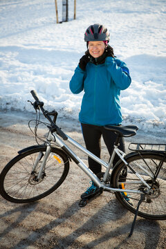 A mid-adult woman puts on a bicycle helmet beside her bike on an icy park path, illustrating safe winter cycling commuting.