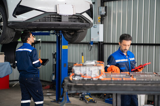 Technician looks up at the underbody of an electric car on a lift while a colleague checks the battery pack nearby.