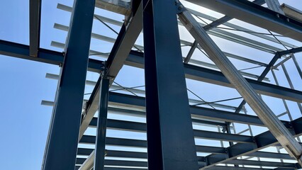 Obraz premium Unfinished steel building frame with blue structural beams, metal roof supports and a wooden ladder against a clear sky at a construction site