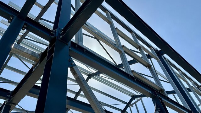 Steel structural framework of an unfinished building with blue beams, galvanized metal supports and bolted connections against a blue sky