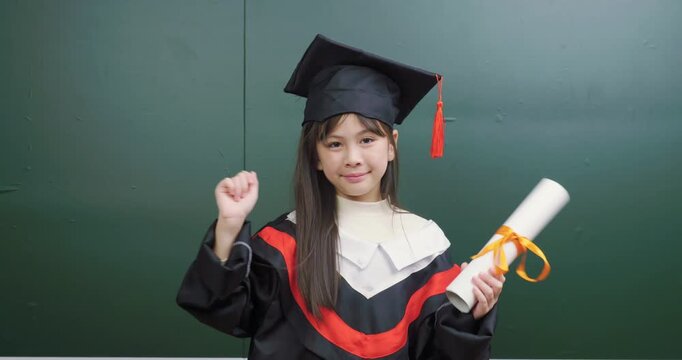 Cute Asian graduate girl holding diploma scroll celebrating success with raised fist gesture.