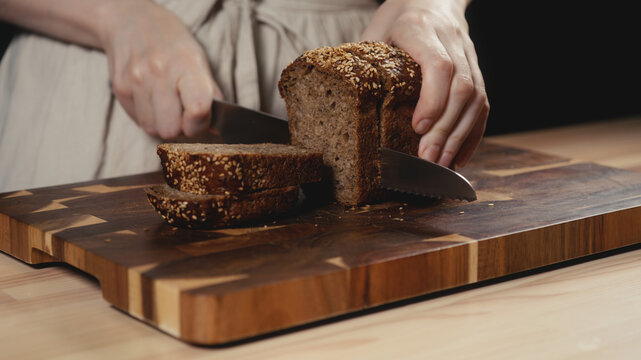 Close-up of baker in rustic linen apron slicing freshly baked artisan whole-grain seed bread with knife on wooden cutting board. Homemade loaf for breakfast, bakery promos, and healthy food concepts.