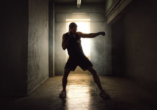 Dramatic silhouette of a strong male boxer throwing a punch with flying dust in a dark underground gym corridor, or intense fighter shadowboxing for competitive martial arts fitness lifestyle.