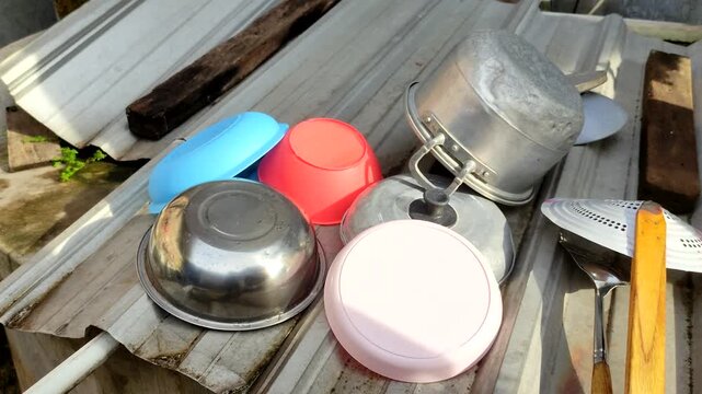 Various colorful plastic bowls, stainless steel pot, lid, and kitchen utensils left drying upside down outdoors on corrugated zinc roofing under bright sunlight.