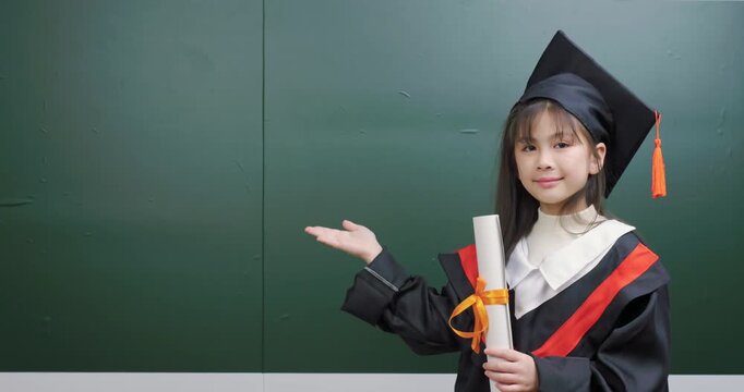 Cute little girl in graduation gown holding diploma certificate and presenting empty space.