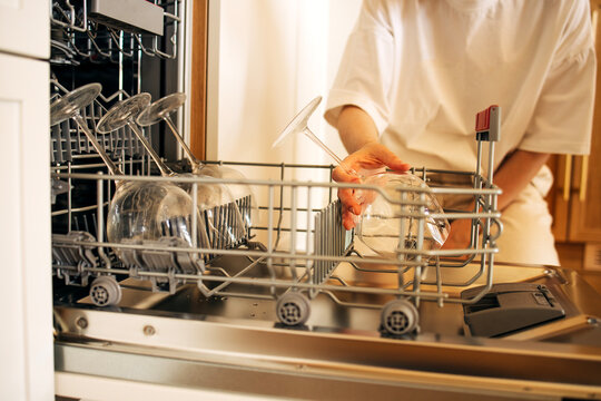 Woman placing clean glassware into a dishwasher rack in a modern kitchen with wooden cabinetry and natural light streaming through the window