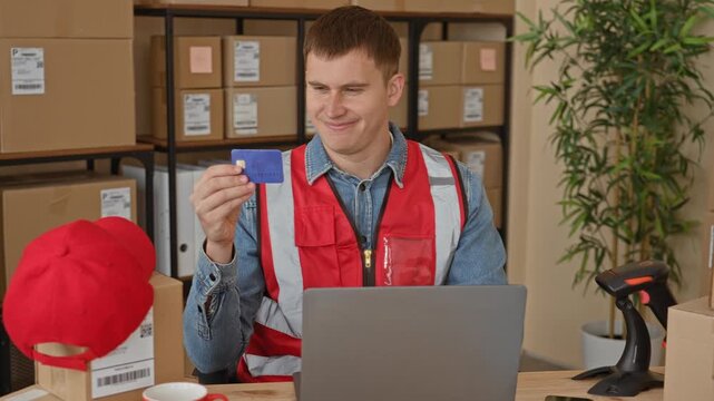 Man courier holding creditcard in hand while smiling at laptop amid stacked packages, cardboard boxes and barcode scanner in a building; satisfaction entrepreneurship.