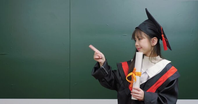 Cute child graduate pointing index finger up and to the side against background.