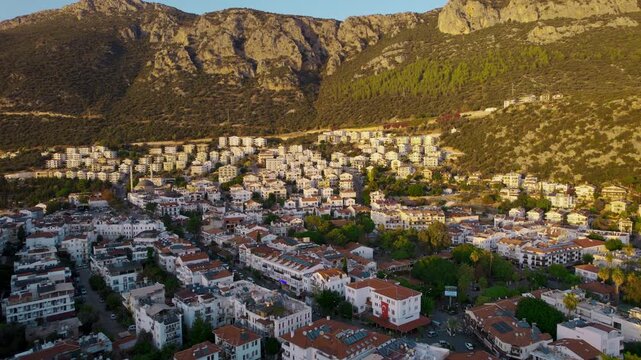 Golden Hour Aerial Over Kas Turkey pass below in warm late day light with white low rise homes red tiled roofs winding streets and a mosque dome and minaret set against steep rocky mountains behind