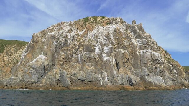 Moving sea level view passes a massive bare granite cliff marked by pale vertical streaks deep fractures and angular ledges while dark blue water ripples below the isolated Primorye headland