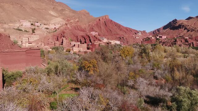 Elevated aerial perspective showcasing traditional red earthen houses nestled directly into the rugged red rock canyon cliffs above the Todra river valley in Morocco.