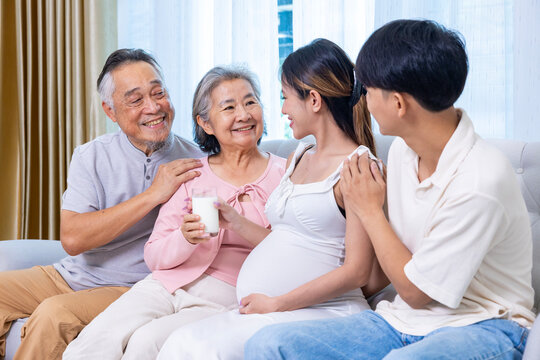 Heavily pregnant asian woman in her third trimester is drinking milk for calcium and nutrition at home with her supporting family and parent in relax and calm for maternity, motherhood, in law concept