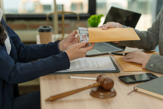 Businessman giving bribe money in envelope to judge or lawyer at desk with wooden gavel and legal documents.