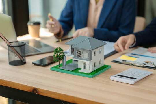 Real estate agent and client discussing house investment plan with laptop and house model on desk.
