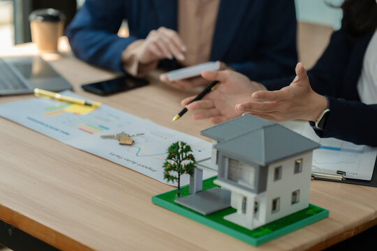 Real estate agent and client discussing house investment plan with laptop and house model on desk.
