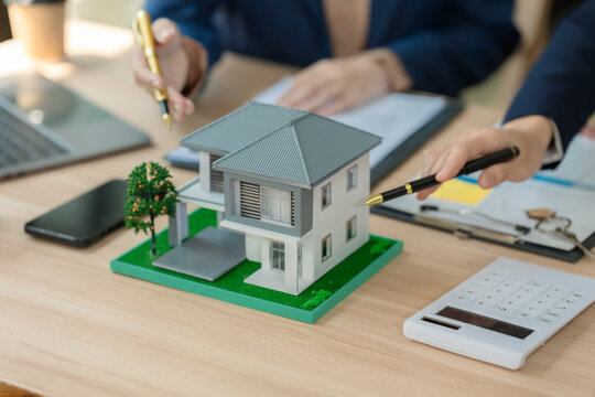 Real estate agent and client discussing house investment plan with laptop and house model on desk.
