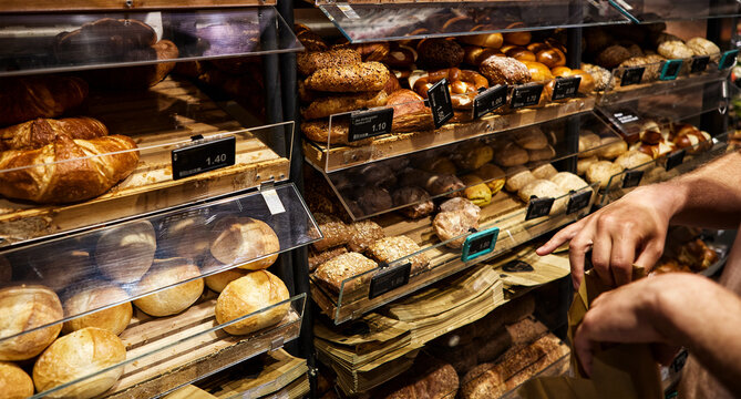 Bakery bread display with assorted loaves, rolls and seeded breads behind transparent shelves, price labels and a customer selecting fresh bread for grocery shopping in a supermarket grocery setting