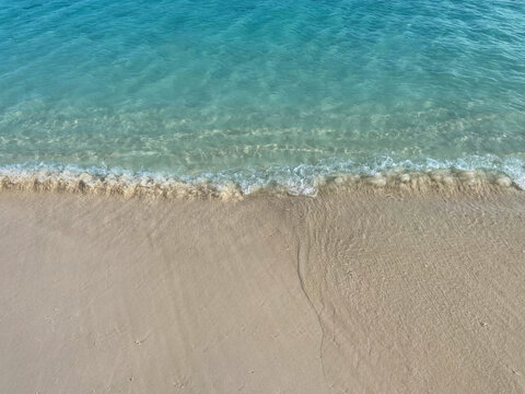 Aerial view of clear turquoise seawater gently meeting a pristine sandy beach, creating a minimalist tropical coastal background.