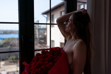 Pensive woman holding red roses stands by a window in an intimate profile portrait © svetograph