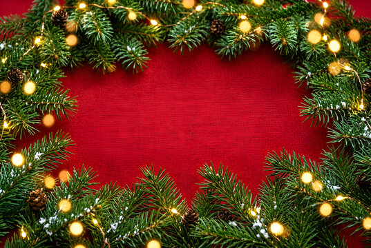 Christmas pine branch border with warm glowing string lights and pinecones on a textured red background. Festive winter holiday frame with central copy space for greeting card template.