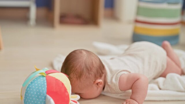 Boy reaching with arm toward colorful soft ball during tummy time inside building; curiosity discovery.