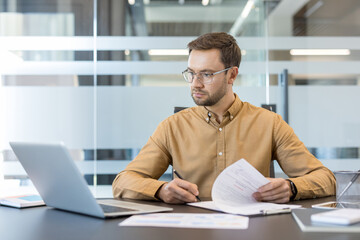 Obraz premium Corporate productivity and business workflow, man working on laptop and reviewing documents in modern office. Professional employee analyzing data and managing tasks with focus and concentration.