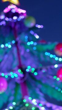 Young woman wearing a yellow beanie looking at an illuminated christmas tree with colorful lights and ornaments at night, with the focus shifting from the person to the treetop star