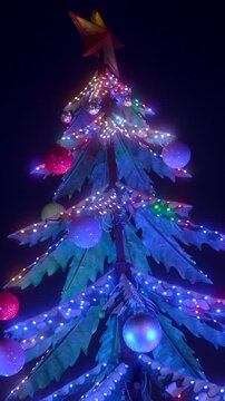Beautifully decorated christmas tree glowing with vibrant, flashing led lights against the dark night sky. The festive holiday installation features bright ornaments, a star, and colorful garlands