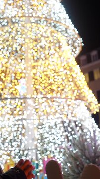 Young woman wearing a yellow beanie admiring a large, brightly lit christmas tree in a city square during the festive winter holidays, enjoying the magical and cheerful evening atmosphere