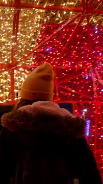 Young woman wearing a yellow beanie and winter jacket marveling at a large, outdoor red and gold christmas light installation, feeling the holiday spirit and excitement in the festive atmosphere