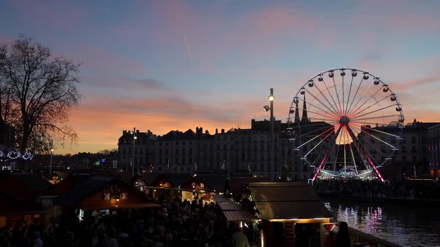 Enchanting christmas market in bayonne, france, glowing at twilight with a large illuminated ferris wheel spinning against a colorful sunset sky reflecting on the nive river