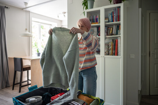 Woman checking clothes for holiday trip before packing things in suitcase at home in bedroom, focused female holding and inspecting sweater before placing in luggage, relaxed vacation preparations.