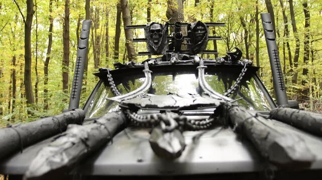 Post-apocalyptic theme, chain details, dark forest. Front close-up showing skull-adorned, heavily armored vehicle with chains, positioned in forest, showcasing grim, survivalist theme.