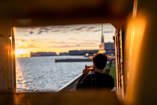 Unrecognizable man filming scene of sunset over bridge and sea bay on smartphone standing on coast, silhouette of man with phone recording golden hour video time lapse of waterfront on mobile camera.