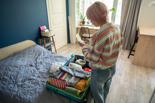 Organized woman traveler marking things from packed suitcase in holiday trip checklist in notepad, pensive female preparing for weekend vacation solo trip and writing off items with pen on paper.