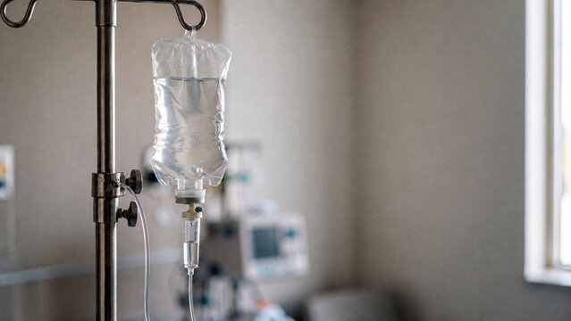Intravenous drip bag hanging on metal stand in hospital room. Blurred medical equipment and bright window in background. Healthcare, infusion therapy and patient treatment concept.