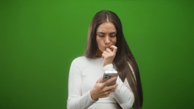 Woman bites fingernail while holding smartphone in studio under green backdrop, furrowed brow and tight mouth; anxiety uncertainty.