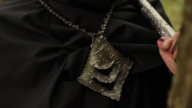 Black cloak, Rugged pendant, Necklace chain. Detailed view presents rugged metal pendant dangling from chain, alongside hand holding weapon handle against black cloak.