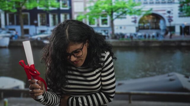 Young brunette woman holding diploma with red ribbon and glasses clutching stomach on street by canal, wincing in pain; graduation setback distress.
