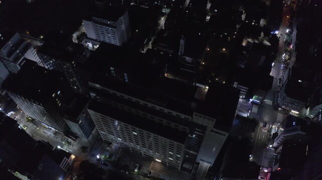 Tall office and apartment blocks form a dark downtown grid as the camera glides overhead with scattered streetlights and traffic below. Seoul