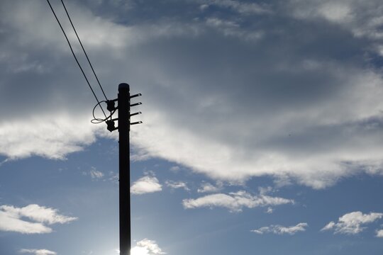 low-angle photograph of a dark utility pole in silhouette, set against a turbulent, heavily overcast sky. The clouds are dark gray and white, suggesting an impending storm or a gloomy day. The power l