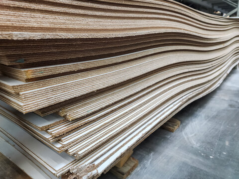 Stacked, curved plywood panels with exposed layered edges showing material quality on a pallet in a warehouse, presenting manufacturing imperfections and a textured background