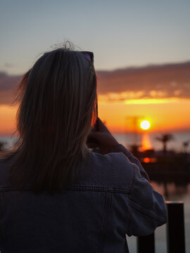 Woman standing with her back to the viewer, capturing a beautiful orange sunset reflecting on the calm ocean surface with palm trees in the distance. Vertical photo