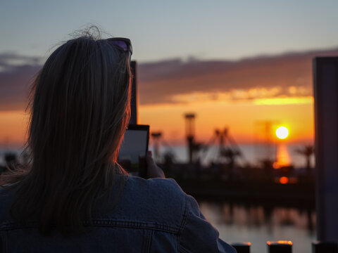 Woman standing on a pier capturing the beautiful orange and yellow sunset view over the tranquil sea with her smartphone during the evening dusk